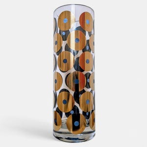 Könnte beinhalten: Eine hohe, transparente Glasvase mit einem sich wiederholenden Muster aus goldenen Kreisen, jeweils mit einem blauen Zentrum. Die Kreise sind mit schwarzen und weißen Formen durchsetzt. Die Vase hat eine einfache, zylindrische Form.