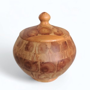 May include: A wooden container with a lid, made of light brown wood with a chequered pattern. The lid has a rounded knob on top.
