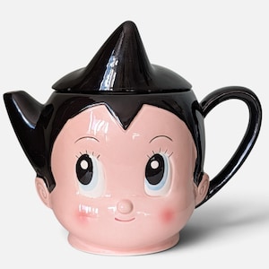 Op de afbeelding: Een keramische theepot in de vorm van het hoofd van Astro Boy, met een glanzend zwart deksel in de vorm van een kapsel met een punt, grote blauwe ogen, roze wangen en een gebogen handvat aan de rechterkant. De theepot heeft een tuit aan de linkerkant.