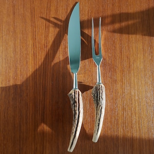 May include: A set of two kitchen utensils with antler handles. The knife has a silver blade and the fork has four tines. The utensils are on a wooden surface.