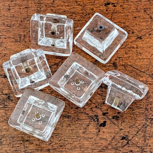 Może przedstawiać: Sześć przezroczystych, kwadratowych szklanych gałek do szafek z mosiężnym osprzętem ułożonych na ciemnej drewnianej powierzchni. Każda gałka ma lekko teksturowany, sześcienny wygląd z centralnym otworem do montażu. Gałki są równomiernie rozmieszczone.