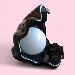 May include: A black ceramic sculpture of an open shell cradling a white sphere. The shell has a glossy finish with wavy edges, and the sphere is smooth and reflective. The sculpture is set against a soft pink background.