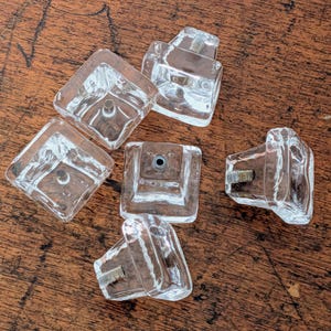 May include: Six clear glass cabinet knobs with a square base and a rectangular top. The knobs are transparent, allowing light to pass through. The hardware is visible within the glass. The knobs are arranged on a wooden surface.