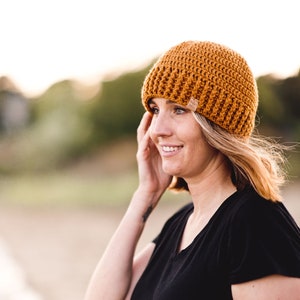 May include: Mustard yellow crochet beanie with a small tag on the side. The hat is made of a textured yarn and is worn by a person wearing a black t-shirt.