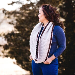 May include: A cream-colored infinity scarf with a dark brown border is worn around the neck. The scarf is made of a knitted material. The person in the image is wearing a blue long-sleeve shirt and blue jeans.