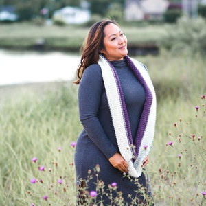 May include: A woman wearing a gray dress and a white and purple crochet infinity scarf. The scarf is draped around the neck. The woman is standing in a field of tall grass and purple flowers.