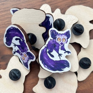 Könnte beinhalten: Ein Satz von acht Holzstiften mit einer lila und weißen Illustration eines Aye-Aye-Lemurs. Die Stifte sind mit schwarzen Metall-Stift-Rückseiten versehen.