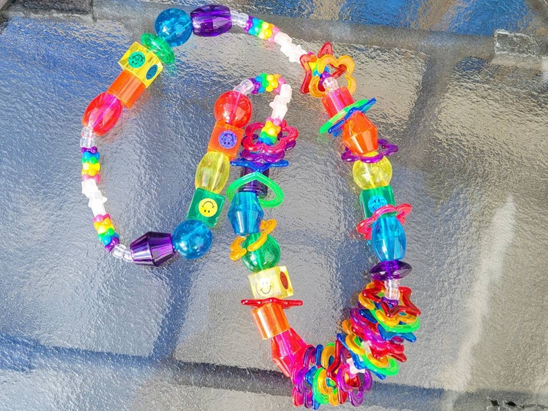 May include: A colorful necklace featuring a variety of plastic beads in shades of red, orange, yellow, green, blue, and purple. The necklace includes star, smiley face, and geometric shaped beads. The necklace is approximately 18 inches long.
