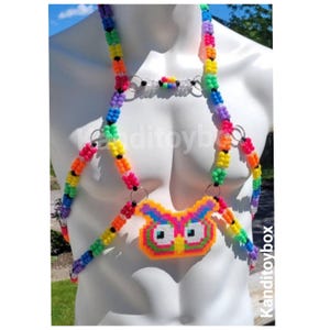 Peut inclure: Un harnais de corps compos&eacute; de perles en plastique multicolores dispos&eacute;es en arc-en-ciel, avec un pendentif central en forme de hibou fait de perles pixelis&eacute;es fusionn&eacute;es. Le design inclut des anneaux m&eacute;talliques argent&eacute;s reliant les brins de perles.