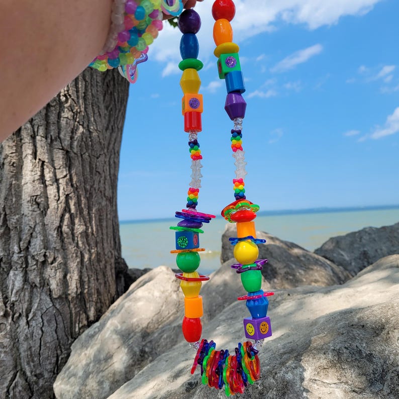May include: A colorful beaded necklace with a variety of shapes and sizes. The necklace features red, orange, yellow, green, blue, and purple beads, with clear beads in between. The necklace is held up against a blue sky and water background.