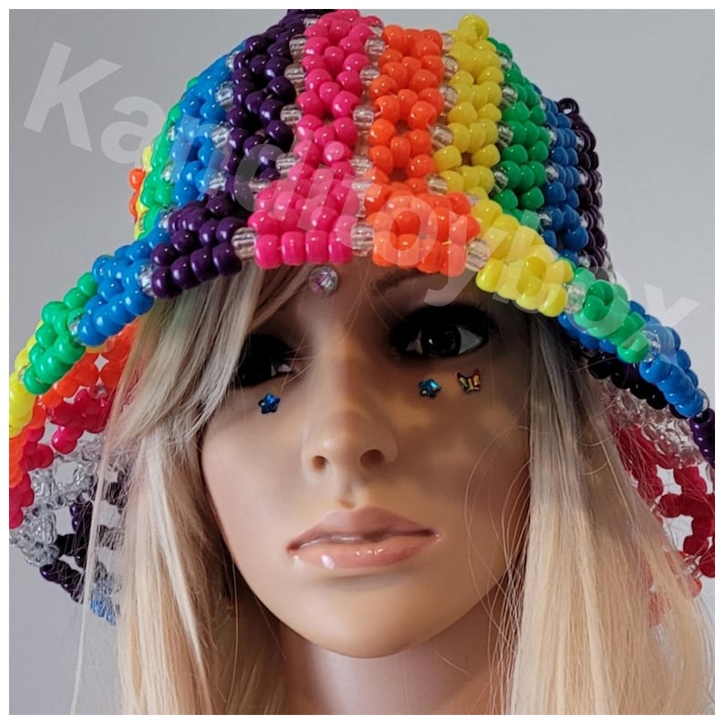 Peut inclure: Un chapeau en perles color&eacute;es avec un motif arc-en-ciel. Le chapeau est compos&eacute; de perles de diff&eacute;rentes couleurs, notamment rouge, orange, jaune, vert, bleu et violet. Le chapeau est port&eacute; par un mannequin aux cheveux blonds et avec des bijoux sur le visage.