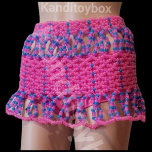 Op de afbeelding: Een rok gemaakt van doorschijnende roze en blauwe plastic kralen in een herhalend rasterpatroon. Het kledingstuk heeft een brede tailleband, een dicht middengedeelte en een geschulpte zoom met bungelende lussen. De tekst luidt Kanditoybox.