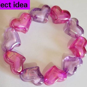 K&ouml;nnte beinhalten: Ein Armband aus herzf&ouml;rmigen Perlen in Rosa- und Lilat&ouml;nen. Die durchscheinenden Perlen sind kreisf&ouml;rmig angeordnet. Der Text "Project idea" befindet sich in einem lila Rechteck oben links.