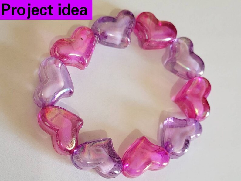 K&ouml;nnte beinhalten: Ein Armband aus herzf&ouml;rmigen Perlen in Rosa- und Lilat&ouml;nen. Die durchscheinenden Perlen sind kreisf&ouml;rmig angeordnet. Der Text "Project idea" befindet sich in einem lila Rechteck oben links.