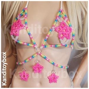 Peut inclure: Un haut de type dos nu composé de perles en plastique multicolores disposées en arc-en-ciel. Le vêtement présente deux grandes étoiles en perles roses sur les bonnets et trois petites étoiles roses suspendues à des anneaux en métal argenté.