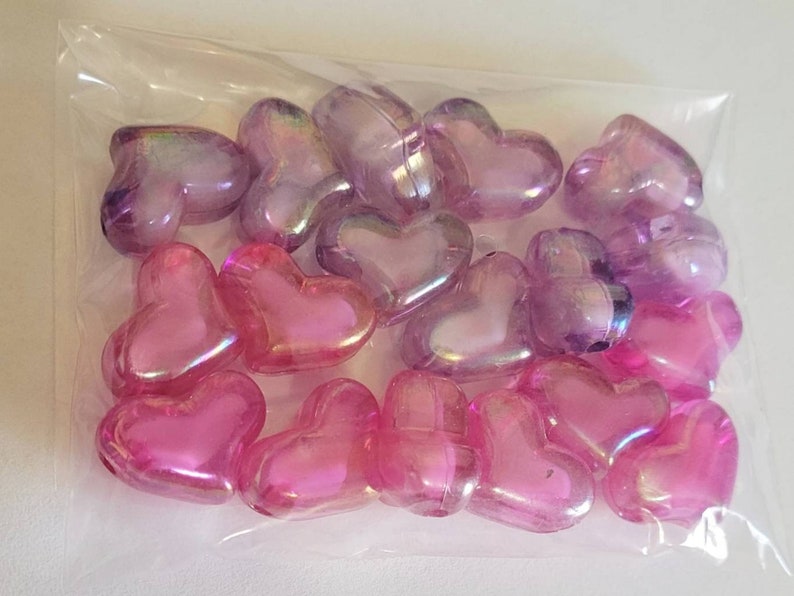K&ouml;nnte beinhalten: Ein klarer Plastikbeutel gef&uuml;llt mit herzf&ouml;rmigen Perlen. Die Perlen sind rosa und lila mit schillerndem Glanz. Die Herzen sind unterschiedlich gro&szlig; und eng beieinander.