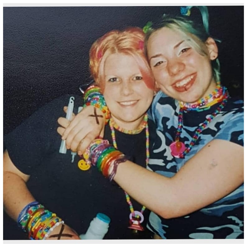 May include: Two smiling people embrace, adorned with colorful beaded necklaces and bracelets. One wears a black shirt with a cross drawn on their arm, while the other sports a blue camouflage top. Both have colorful hair and accessories.