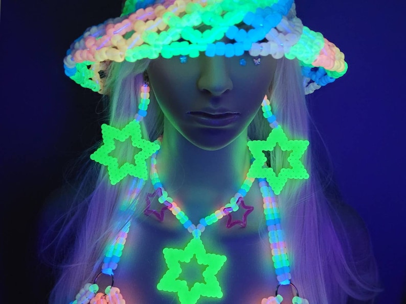 May include: A brightly glowing rave outfit, featuring a beaded hat, earrings, and a necklace with star pendants. The accessories glow against a dark backdrop, creating a vibrant, eye-catching look. The outfit is made of colourful beads.