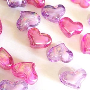 K&ouml;nnte beinhalten: Eine Sammlung durchscheinender herzf&ouml;rmiger Perlen in Rosa- und Lilat&ouml;nen. Die Perlen haben eine gl&auml;nzende Oberfl&auml;che und sind auf einer wei&szlig;en Oberfl&auml;che verstreut, was eine romantische und skurrile &Auml;sthetik erzeugt. Ideal f&uuml;r Bastelarbeiten oder die Schmuckherstellung.