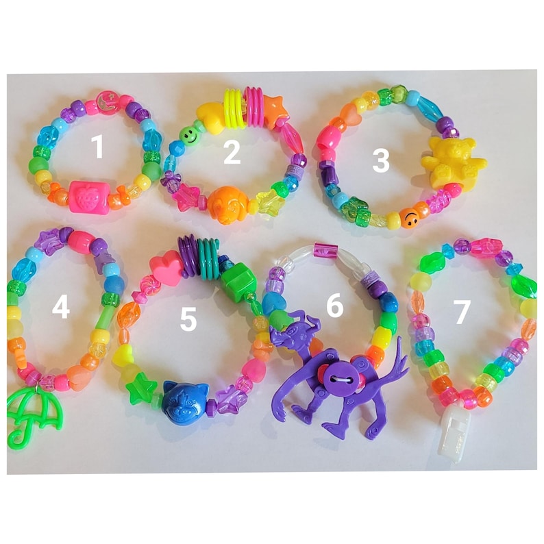 May include: Seven colorful beaded bracelets with various charms, including a pink square, a yellow star, a yellow bear, a green umbrella, a blue cat, a purple monkey, and a white rectangle. The bracelets feature rainbow-colored beads.