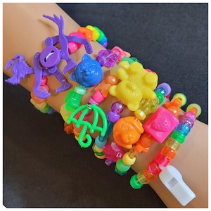 May include: A collection of colorful beaded bracelets with various plastic charms, including a purple robot, a blue cat, a yellow bear, and a green umbrella. The bracelets feature rainbow-colored beads and a white whistle.