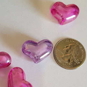 K&ouml;nnte beinhalten: Mehrere durchscheinende herzf&ouml;rmige Perlen in Rosa- und Lilat&ouml;nen liegen auf einer wei&szlig;en Oberfl&auml;che. Eine US-Viertel-Dollar-M&uuml;nze dient als Gr&ouml;&szlig;envergleich. Die Perlen haben eine gl&auml;nzende Oberfl&auml;che.