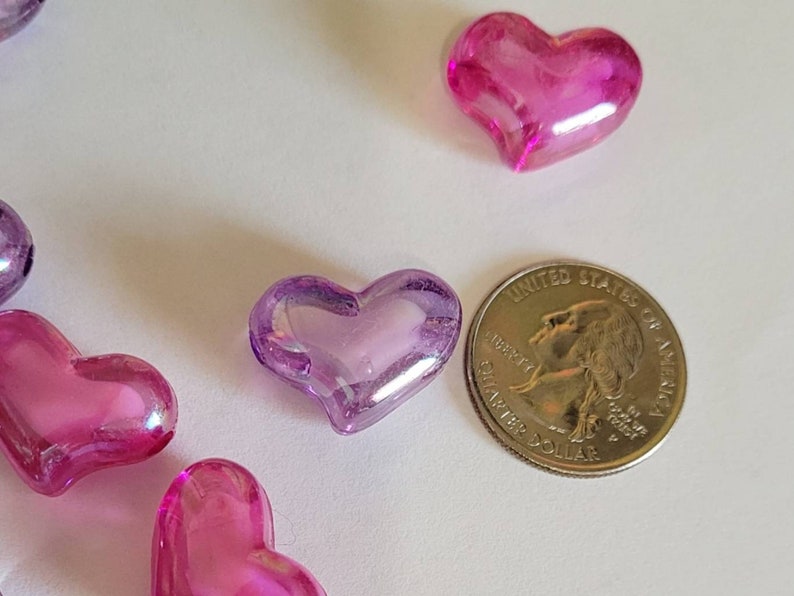 K&ouml;nnte beinhalten: Mehrere durchscheinende herzf&ouml;rmige Perlen in Rosa- und Lilat&ouml;nen liegen auf einer wei&szlig;en Oberfl&auml;che. Eine US-Viertel-Dollar-M&uuml;nze dient als Gr&ouml;&szlig;envergleich. Die Perlen haben eine gl&auml;nzende Oberfl&auml;che.