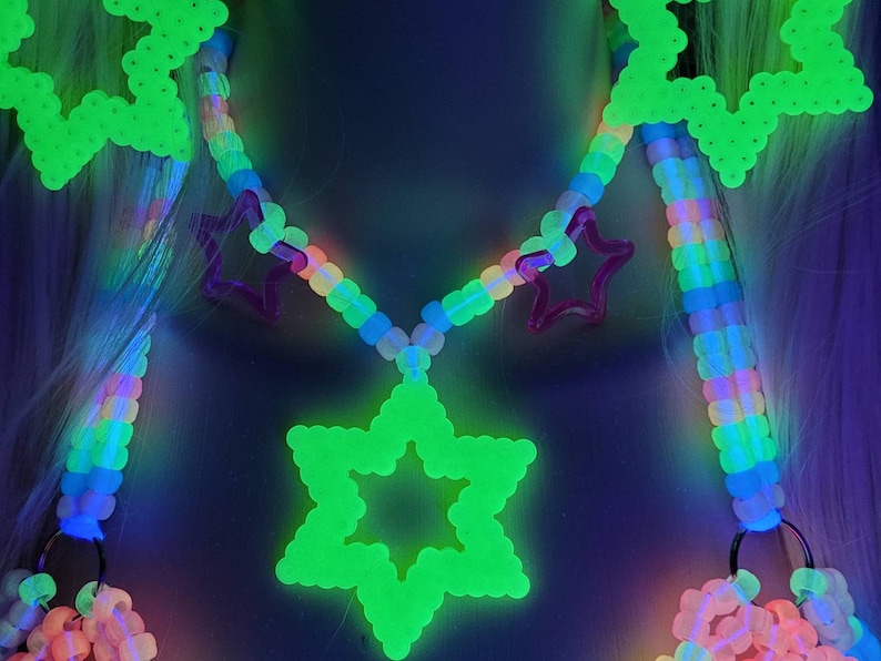 May include: A neon green star pendant necklace with a rainbow beaded chain and pink star accents. The necklace glows brightly against a dark background. The design includes a large star pendant and smaller star shapes.