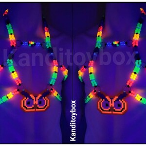 Peut inclure: Un harnais de corps en perles aux couleurs n&eacute;on avec un pendentif hibou pix&eacute;lis&eacute;. Le harnais est compos&eacute; de perles en plastique vert, jaune et orange vif reli&eacute;es par des anneaux m&eacute;talliques, brillant sous lumi&egrave;re UV avec le texte Kanditoybox.