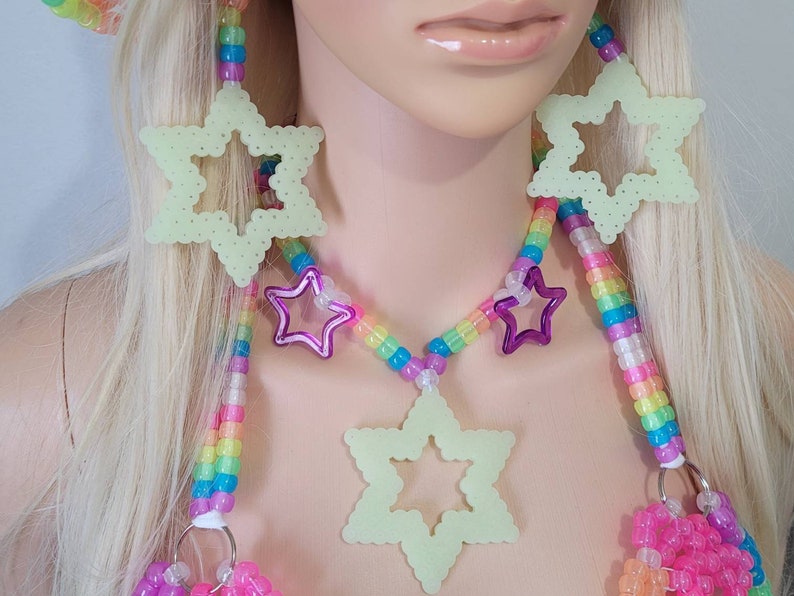 May include: A neon green star-shaped necklace and earrings set with a rainbow beaded chain. The necklace features a large star pendant and smaller purple star accents. The earrings are also star-shaped. The set is made of plastic beads.