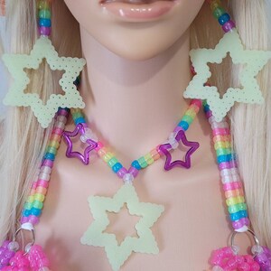 May include: A vibrant rave-style necklace and top set. The necklace has a rainbow beaded design with purple star accents and large, glowing star pendants. The top is made of pink and purple beads.