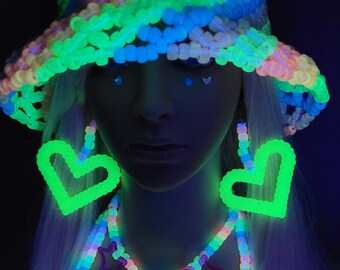 May include: A neon bucket hat, heart-shaped earrings, and a beaded necklace made of multi-colored plastic pony beads. The accessories glow in a vibrant, luminous green and blue under ultraviolet light, creating a high-contrast effect.