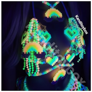Pu&ograve; includere: Un set di reggiseno e accessori di perline fatti a mano di colore neon. Il reggiseno presenta un design a cuore e stella, con accenti arcobaleno e nuvole. Il set si illumina al buio, con il testo "Kanditoybox" nell'angolo.