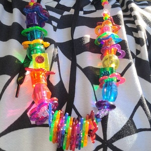 May include: A colorful, beaded necklace with a variety of transparent plastic shapes, including flowers, hearts, and cylinders. The necklace features a smiley face charm and a rainbow-colored section. The beads are strung on a clear cord.