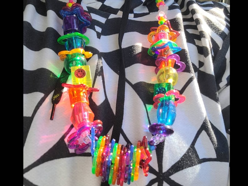 May include: A colorful, beaded necklace with a variety of transparent plastic shapes, including flowers, hearts, and cylinders. The necklace features a smiley face charm and a rainbow-colored section. The beads are strung on a clear cord.