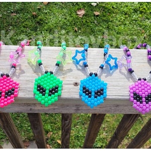 May include: Four beaded necklaces featuring alien-shaped pendants made of small plastic beads in neon pink, bright green, cyan blue, and purple. Each necklace includes clear and black accent beads with star-shaped charms attached to the strands.