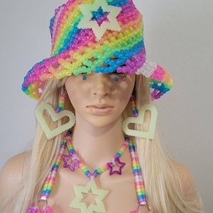 May include: A rainbow-coloured beaded bucket hat with a star accent, matching earrings, and a necklace with star and heart pendants. The accessories are neon green and the hat is made of colourful beads.