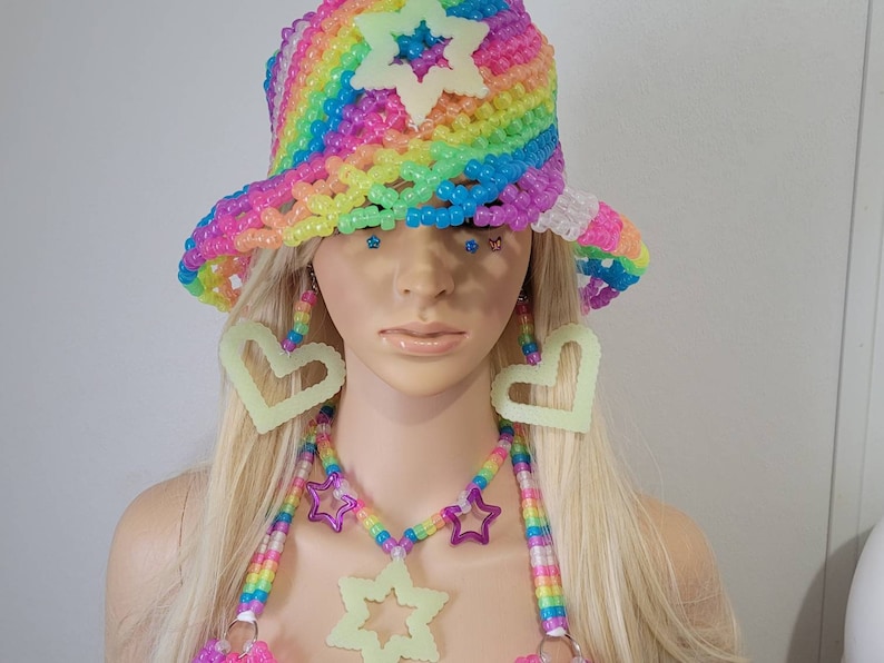 May include: A rainbow-coloured beaded bucket hat with a star accent, matching earrings, and a necklace with star and heart pendants. The accessories are neon green and the hat is made of colourful beads.