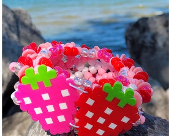 May include: A colorful beaded bracelet with a strawberry design. The bracelet features pink, red, and white beads, with two strawberry charms in pink and red. The strawberries have green tops and white square details. The bracelet is on a rock with a blue ocean background.