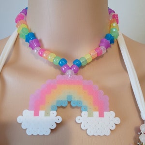Peut inclure: Un collier de perles colorées avec un pendentif arc-en-ciel et des perles en forme d'étoile. Le pendentif arc-en-ciel est fait de perles carrées aux couleurs pastel, avec des nuages blancs. Le collier est présenté sur un mannequin.