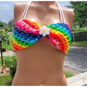 Peut inclure: Un haut de bikini style dos nu fabriqué à partir de perles en plastique multicolores disposées en arc-en-ciel. Le vêtement présente des liens blancs et une décoration en forme de fleur en plastique blanc au centre de l'encolure.