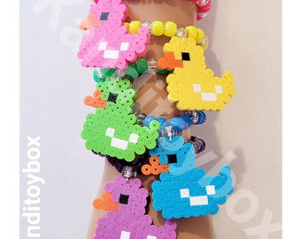 May include: A stack of five colorful beaded bracelets, each featuring a pixelated duck charm made from plastic fuse beads. The bracelets use vibrant pony beads in pink, yellow, green, blue, and purple, accented with clear beads and orange beaks.