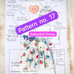 Può includere: Un vestito per bambola bianco con un motivo ripetuto di rose rosse, figure vestite di blu e cuori rossi, dotato di maniche corte e un bordo in pizzo bianco sull'orlo, posizionato sopra un cartamodello con l'etichetta Pattern no. 17.