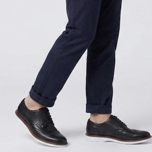 May include: A pair of black leather oxford shoes with decorative brogue detailing, thin black laces, and a thick white rubber sole. The shoes are worn with dark navy blue trousers that are cuffed at the bottom, showing a clean and simple design.