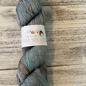 May include: A skein of yarn in shades of blue, gray, and brown. The yarn is wrapped with a white label that reads "Yarn Indulgence" and features a small illustration. The yarn is displayed against a light wood background.