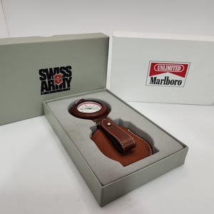 May include: A Swiss Army brand pocket watch with a brown leather case and strap, presented in a grey box. The box lid features the Swiss Army logo, while a separate white box displays the Marlboro Unlimited logo in red, white, and black.