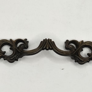 Könnte beinhalten: Bronzefarbener Schrankgriff mit aufwendigem, sich windendem Blumendesign. Der Griff ist gebogen mit dekorativen Verzierungen an jedem Ende. Ein klassisches Hardware-Teil für Möbelrestaurierung.