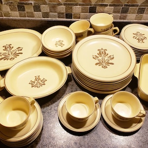 Op de afbeelding: Een vintage set van geel en bruin aardewerk servies met een bloemenpatroon. De set bevat borden, kommen, kopjes, schoteltjes, een botervloot, zout- en peperstrooiers en een ovenschaal.