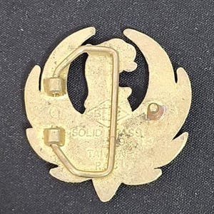 May include: A gold-colored belt buckle with a stylized bird design. The buckle has a rectangular frame and a pin for securing the belt. The text "SOLID BRASS TAIWAN R.O.C." is visible on the buckle.