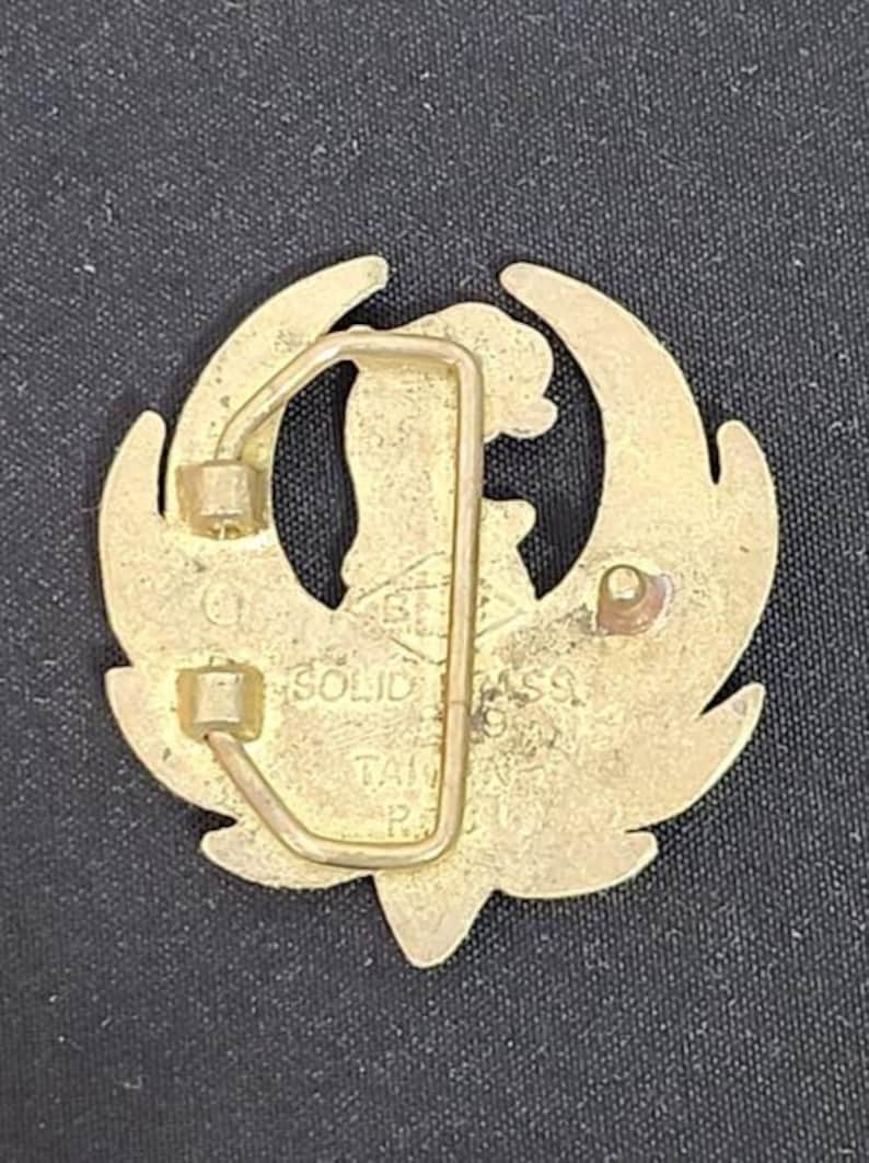 May include: A gold-colored belt buckle with a stylized bird design. The buckle has a rectangular frame and a pin for securing the belt. The text "SOLID BRASS TAIWAN R.O.C." is visible on the buckle.